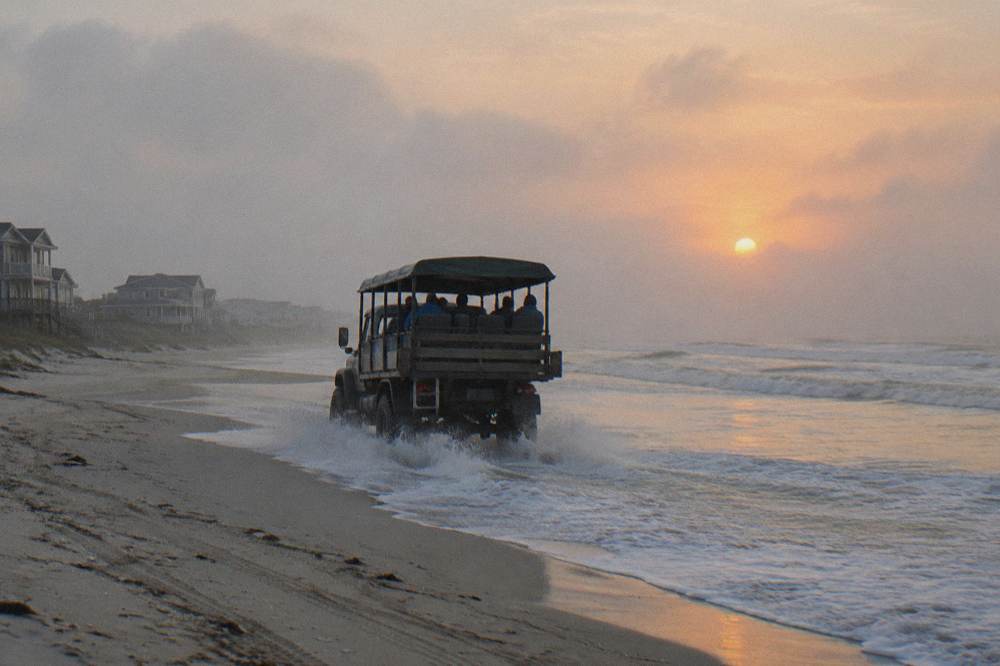 An open-air off-road truck driving along a wide sandy beach at the water line