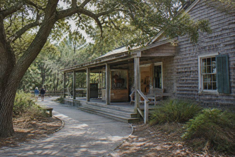 A weathered wooden museum building surrounded by maritime forest near the Corolla village road