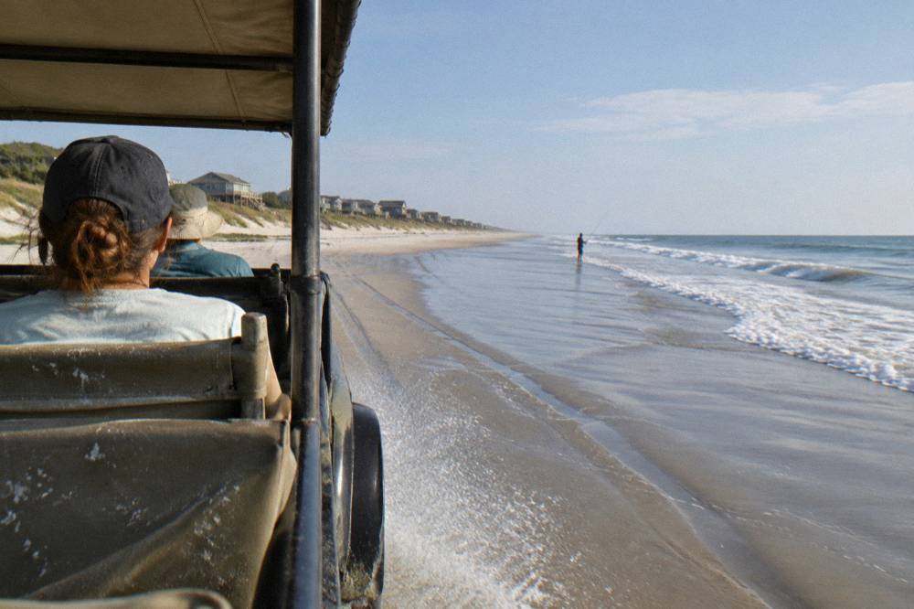 An open-air four-wheel-drive truck driving along a wide sandy beach at low tide.