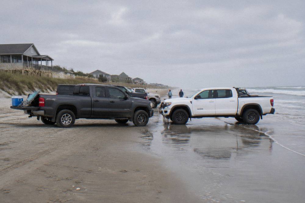 Four-wheel-drive trucks parked on a wide wet-sand beach beside low dunes