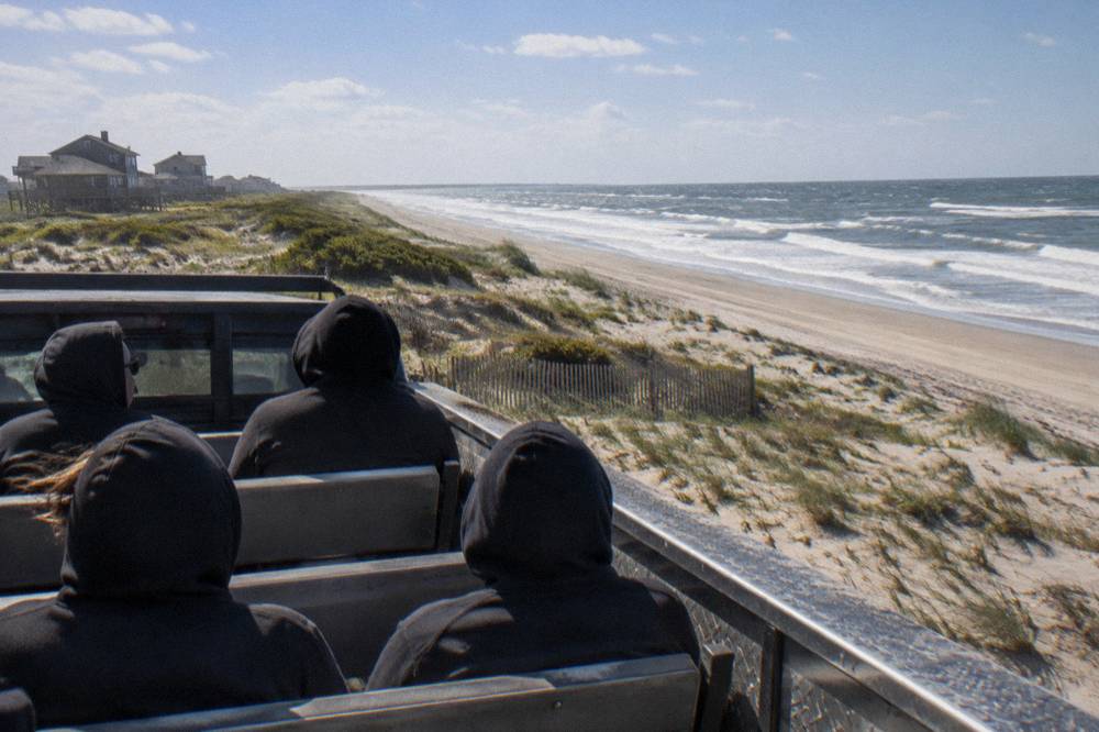 Small group of passengers seated in the open back of a beach tour truck facing the dunes