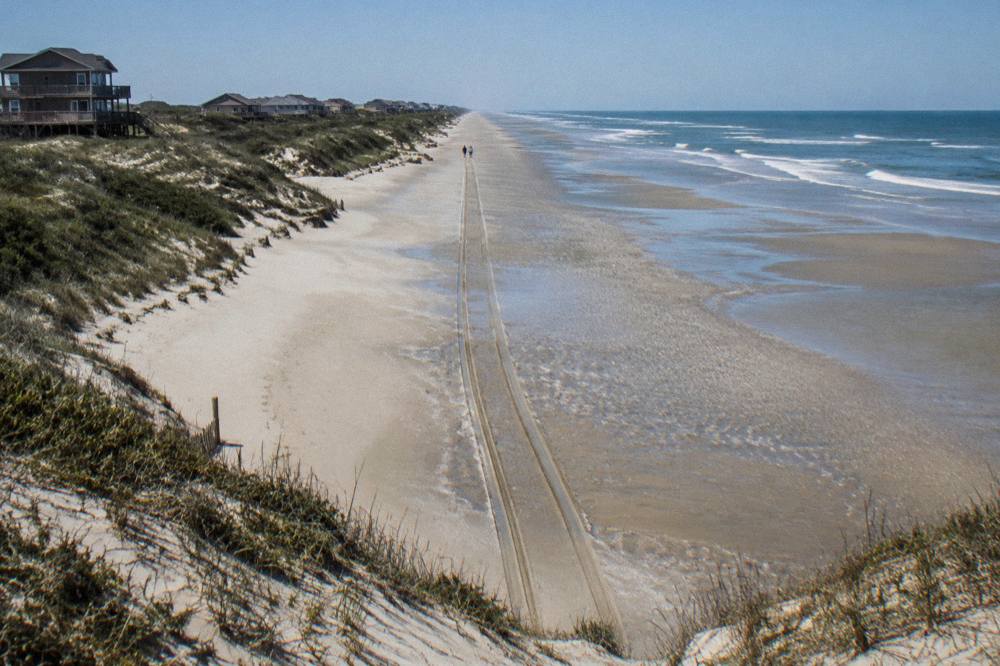 Sand beach road at low tide with a line of tire tracks running north toward empty dunes