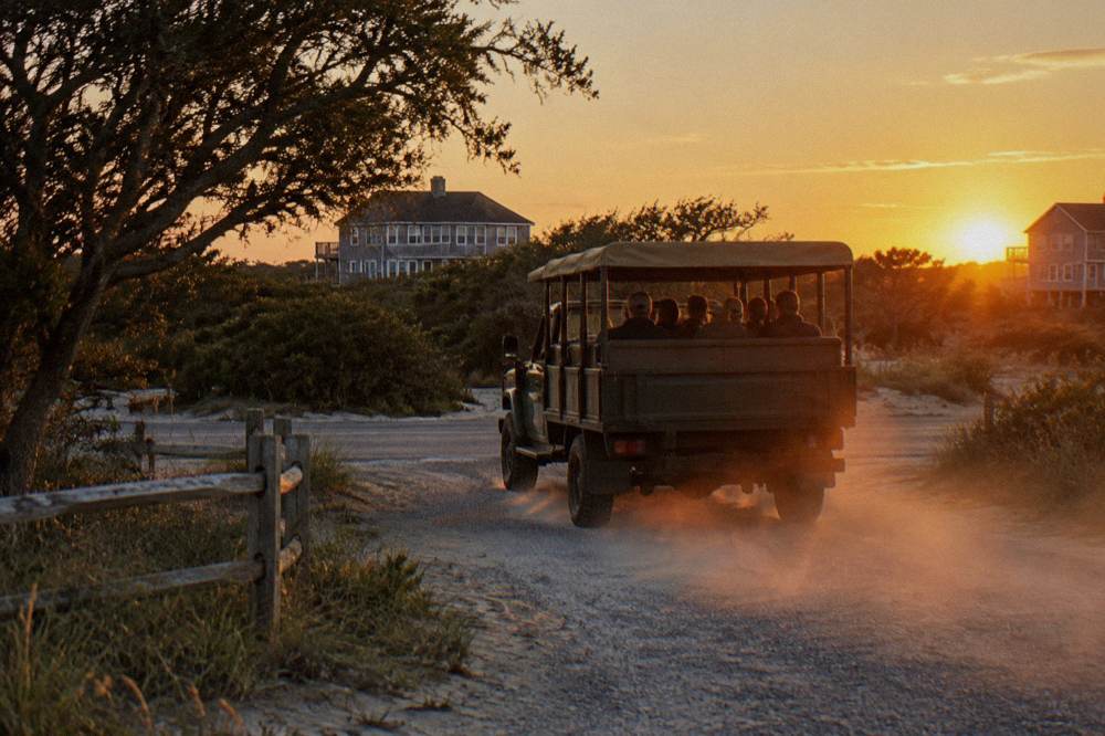 An open-air four-wheel-drive tour truck driving along a wide damp beach at sunset