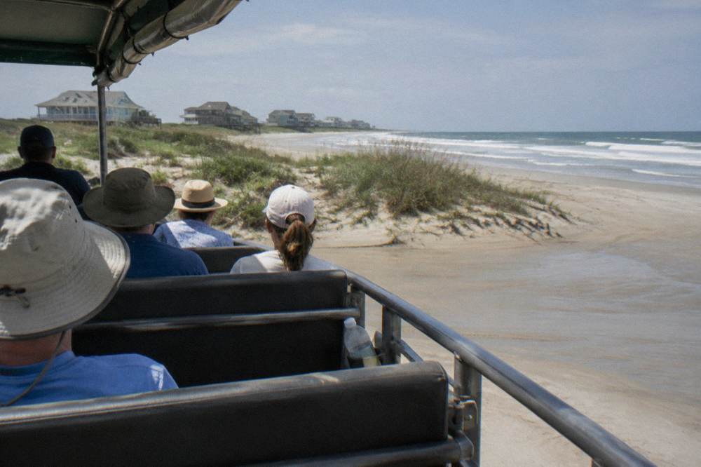 Tour truck with a small group of seated passengers parked on the beach beside dune grass