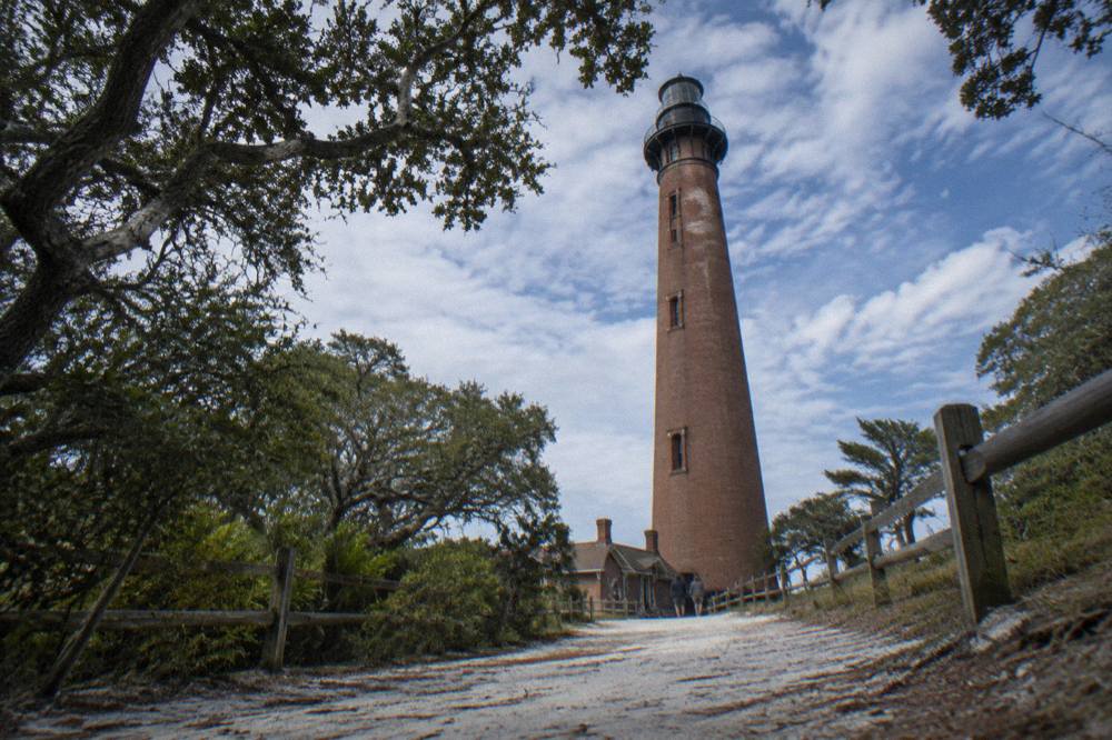 Red and white brick lighthouse tower rising above trees beside a gravel path
