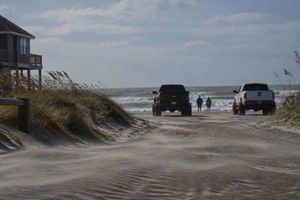 Four-wheel-drive trucks parked on a wide wet-sand beach with the ocean on one side and tall grassy dunes on the other